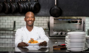 a smiling african american chef proudly displays a plated gourmet dish in a restaurant kitchen